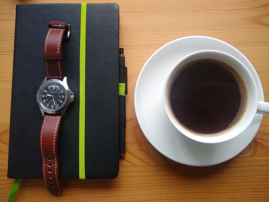 A Hamilton watch resting on a black notepad, beside a cup of black coffee